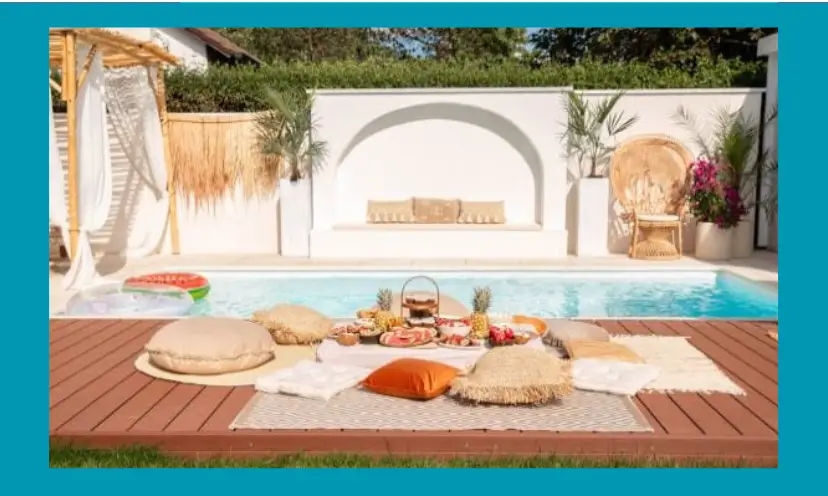 In this photo there is a sparkling pool beside a wooden boardwalk type space for a picnic. In fact, there is an array of food and snacks among comfy pillows with additional rattan seating on the other side of the pool.