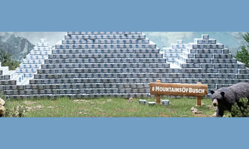 This photo features mountains formed by stacked cans of Busch beer. There are real mountains also in the background. In the forefront of the photo is a wooden sign that says "#Mountains of Busch" and beside the sign is a black bear.