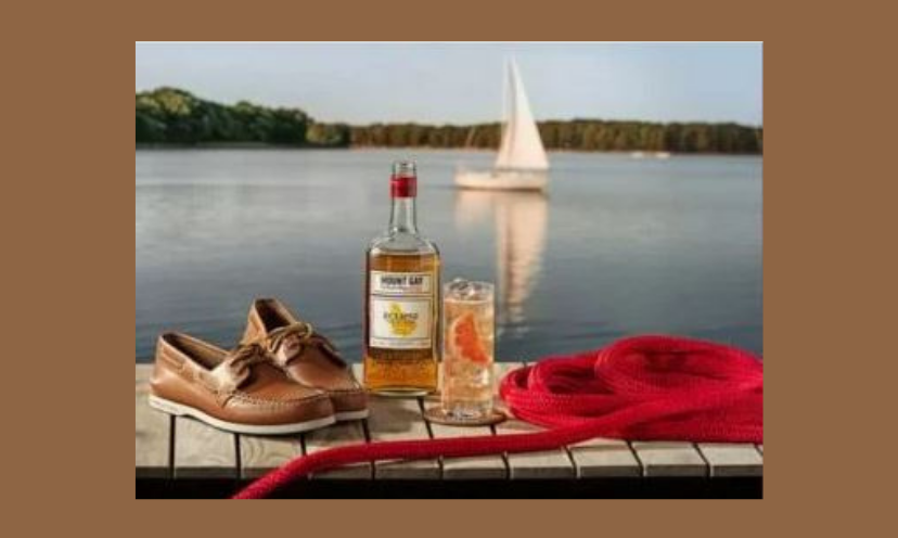 This photo shows one pair of the stylish boat shoes in this giveaway as they share the view with a sailboat on a lake, a bottle of rum and a touch of red with a nautical rope.