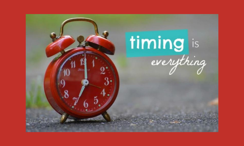 This is a photo of a red alarm clock sitting on a road with the caption "timing is everything"