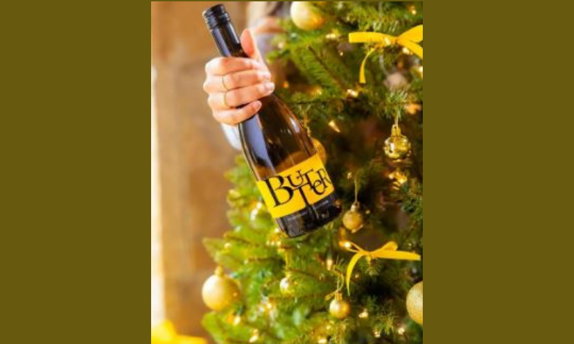 This is a photo of a bottle of Butter Wine being held in front of a Christmas tree decorated with colors matching the bottle itself...golden yellows and green.