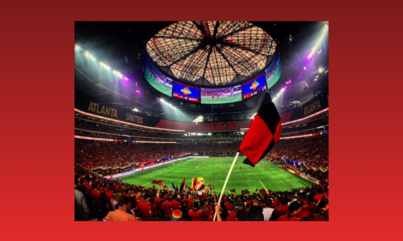 This is a colorful photo of an Atlanta United soccer game in progress. It includes the field, players and excited fans with team supportive flags waving them on!