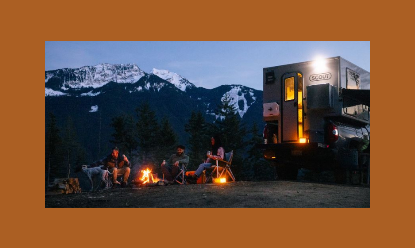 This photo features the Scout camper parked in a cozy setting with snow capped mountains as a backdrop and a blazing campfire warming the people and pups gathered around it.