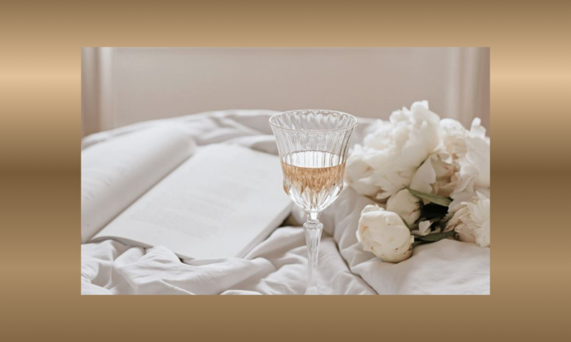 This is a picture of luxury featuring plush bedding, fresh flowers, a comforting drink in a fancy glass and a book waiting to be read!